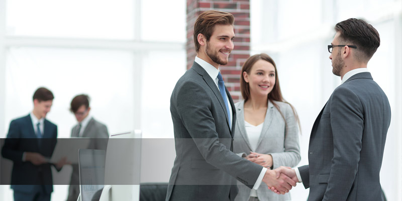Bürokulisse mit weißen Fenstern und einem Ziegelsteinbalken als Kontrast. Im Hintergrund stehen, leicht verschwommen, zwei Männer in Anzügen. Im Vordergrund schütteln zwei Männer einander die Hände. Neben einem der beiden Männer steht eine junge Frau und lächelt.