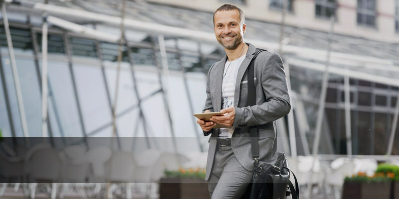 Reinisch Karriere Flexibilität Ein Mann in einem grauen Anzug und mit einer Umhängetasche hält ein kleines Tablet und lächelt dem Betrachter zu