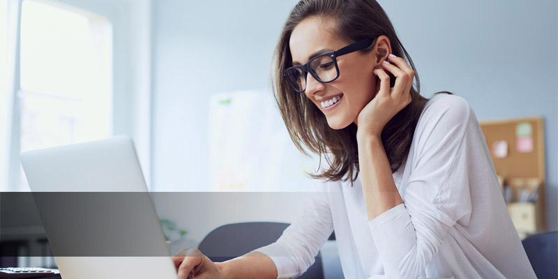 Reinisch Karriere Homeoffice Eine Frau in einer Bürokulisse lächelt einem Laptop zu uns streicht sich dabei die langen braunen Haare hinter das Ohr