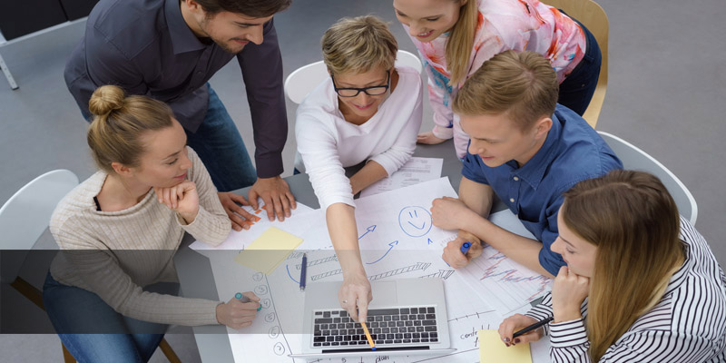 Reinisch Karriere Verantwortungsvolles Arbeiten Um einen Tisch versammelt arbeitet eine Gruppe aus sieben Personen an einem Laptop. Auf dem Tisch liegen mehrere Notizzettel und Stifte.