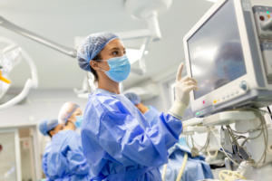 Eine Chirurgin in OP-Kleidung und Gesichtsmaske bedient einen medizinischen Bildschirm im Operationssaal, während im Hintergrund weitere medizinische Fachkräfte bei der Arbeit zu sehen sind.