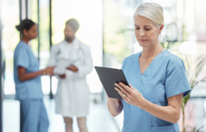 Eine Frau in einem hellblauen Krankenhauskittel konzentriert sich auf das Lesen eines Tablets, während im unscharfen Hintergrund eine weitere Pflegekraft mit einem Arzt kommuniziert.